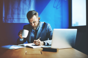 Man drinking coffee as he takes notes on the skills that employers are looking for while searching on his laptop.