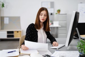 businesswoman going through her papers and working on her desk in front of her screen