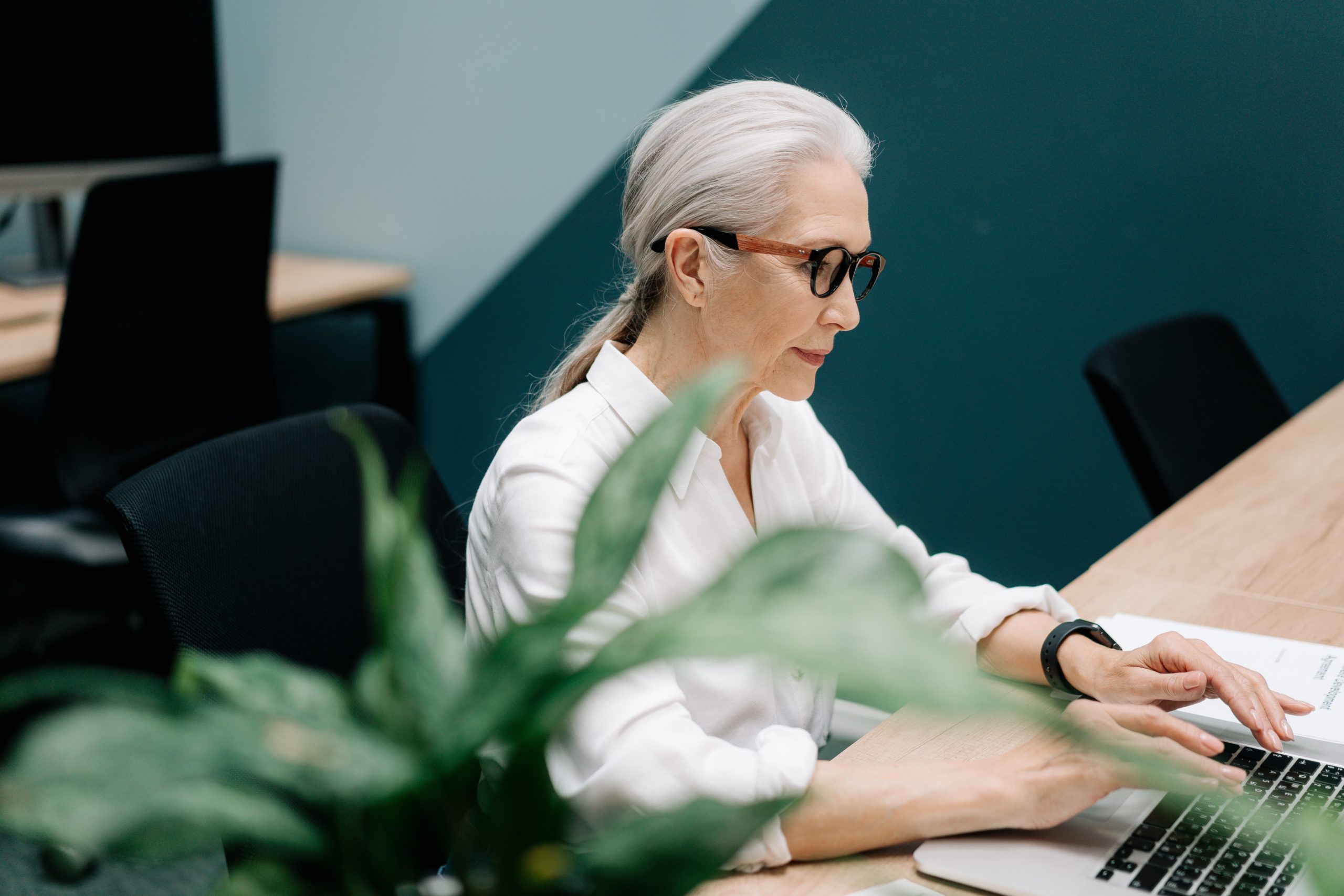 Woman on her 50s wearing a white long sleeves, and eye glasses working on her laptop in the age of ageism