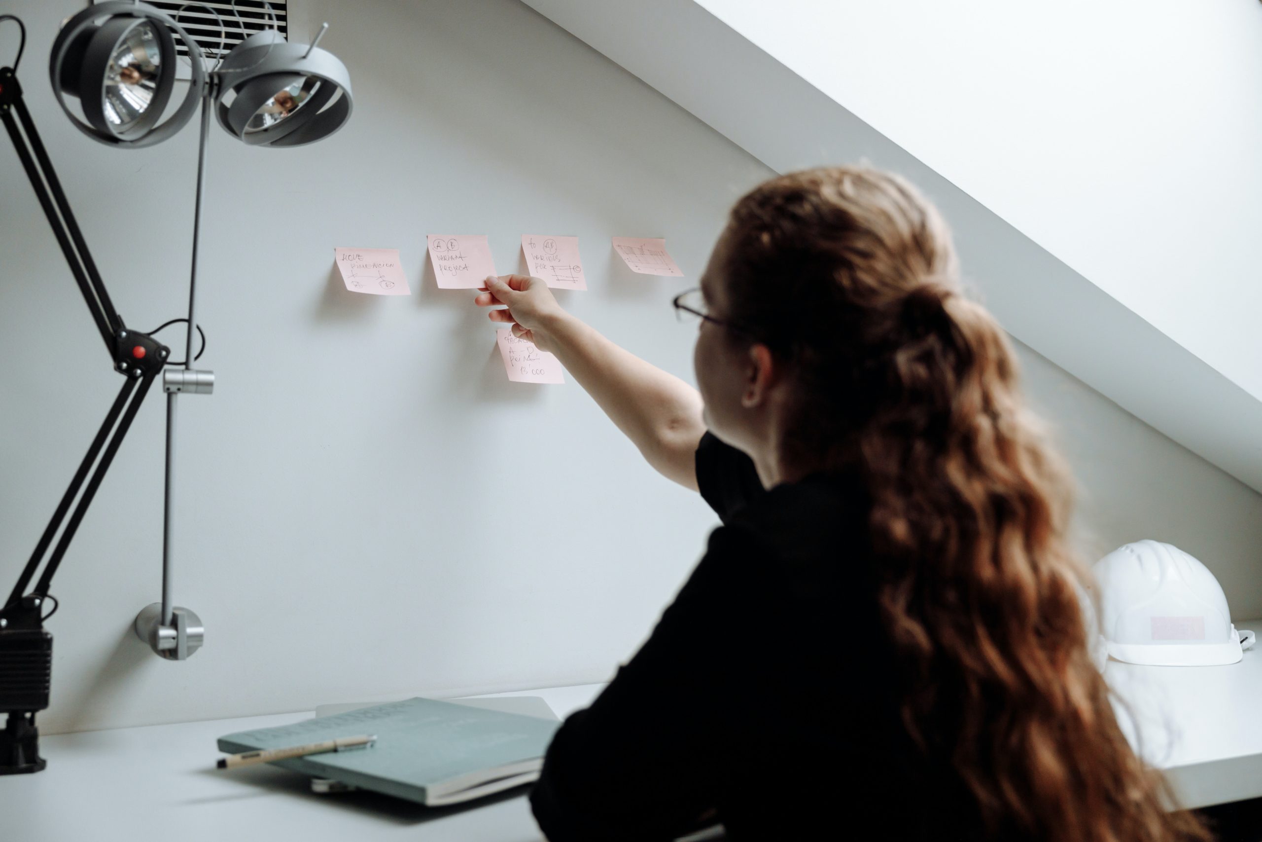 a girl with long curly hair and wearing eye glasses is sitting on her desk at home putting sticky notes on her wall to be a productive a remote worker